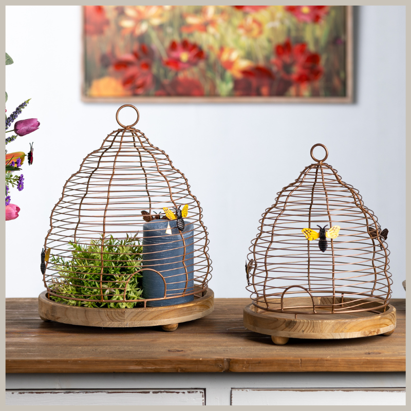A set of bee hive stands with round wooden bases on legs with wired hives, adorned with decorative bees. Place candles or greenery inside. Sitting on a wooden counter. 