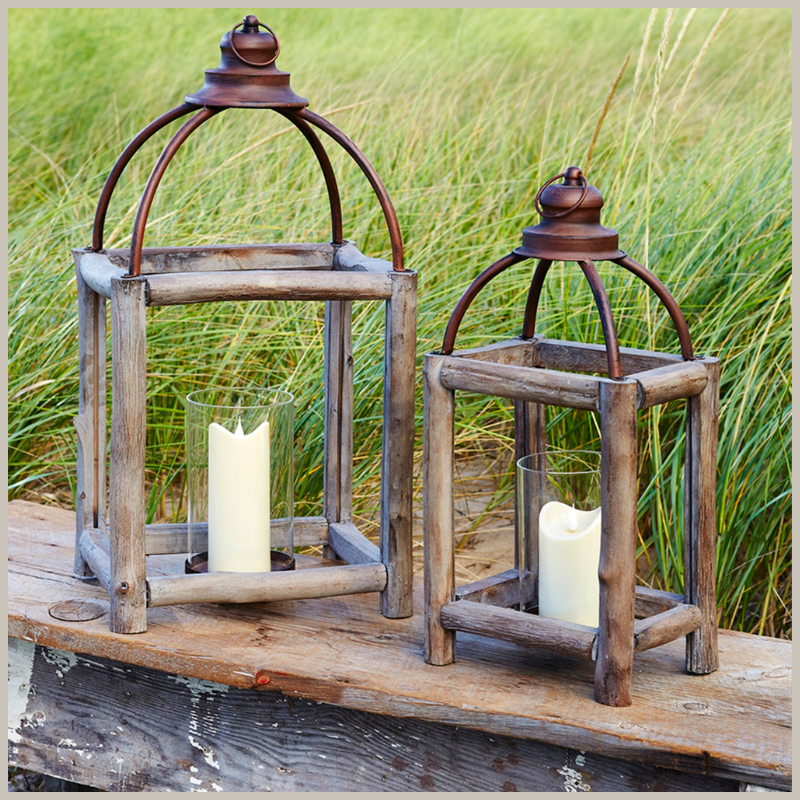 A set of distressed wood branch lanterns with open arched iron toppers. Place your real wax or LED candles in the hurricane candle holder. Sitting on a wooden bench with grasses in background. 