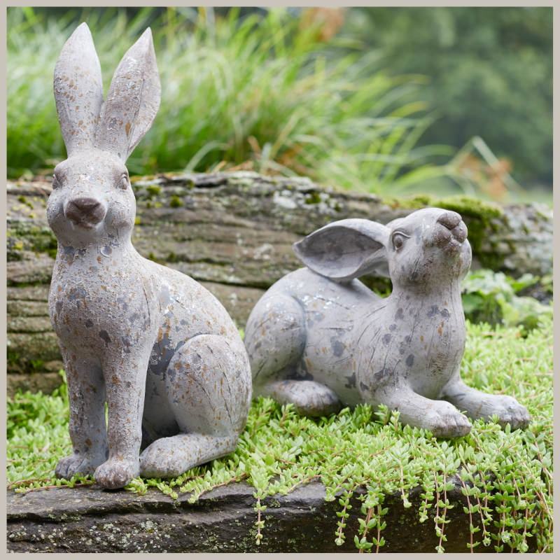A pair of expressive rabbits in weathered stone. One is sitting, the other laying down. Hues of grey and tan. Shown in a garden. 